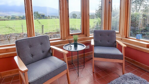 Una habitación con dos sillones grises, una mesita redonda con una planta en maceta y ventanas amplias que dan a un campo verde y montañas distantes.
