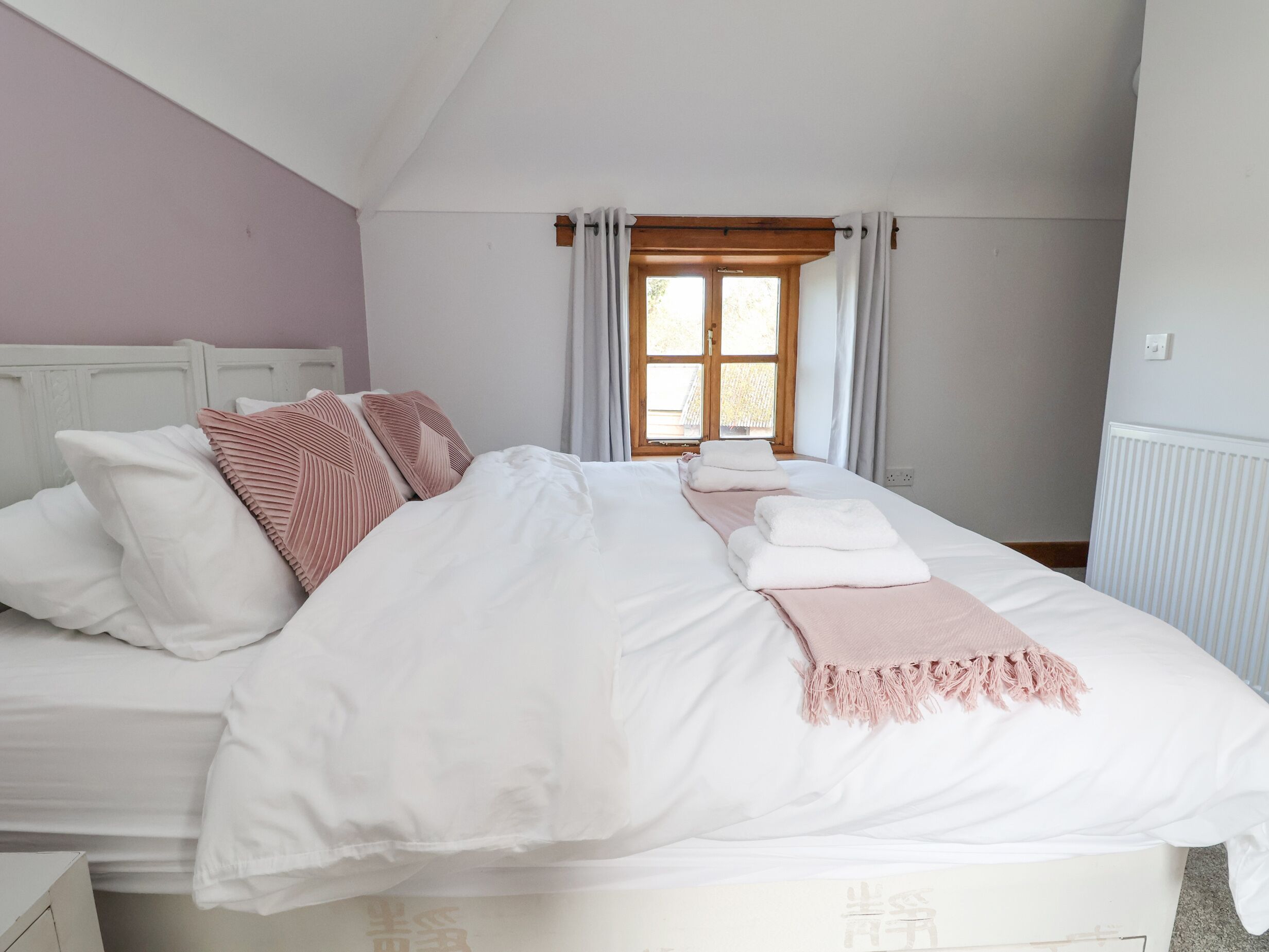 Uma cama bem arrumada com roupa de cama branca e rosa, uma cabeceira de madeira e uma janela com cortinas.