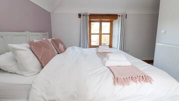 Uma cama bem arrumada com roupa de cama branca e rosa, uma cabeceira de madeira e uma janela com cortinas.
