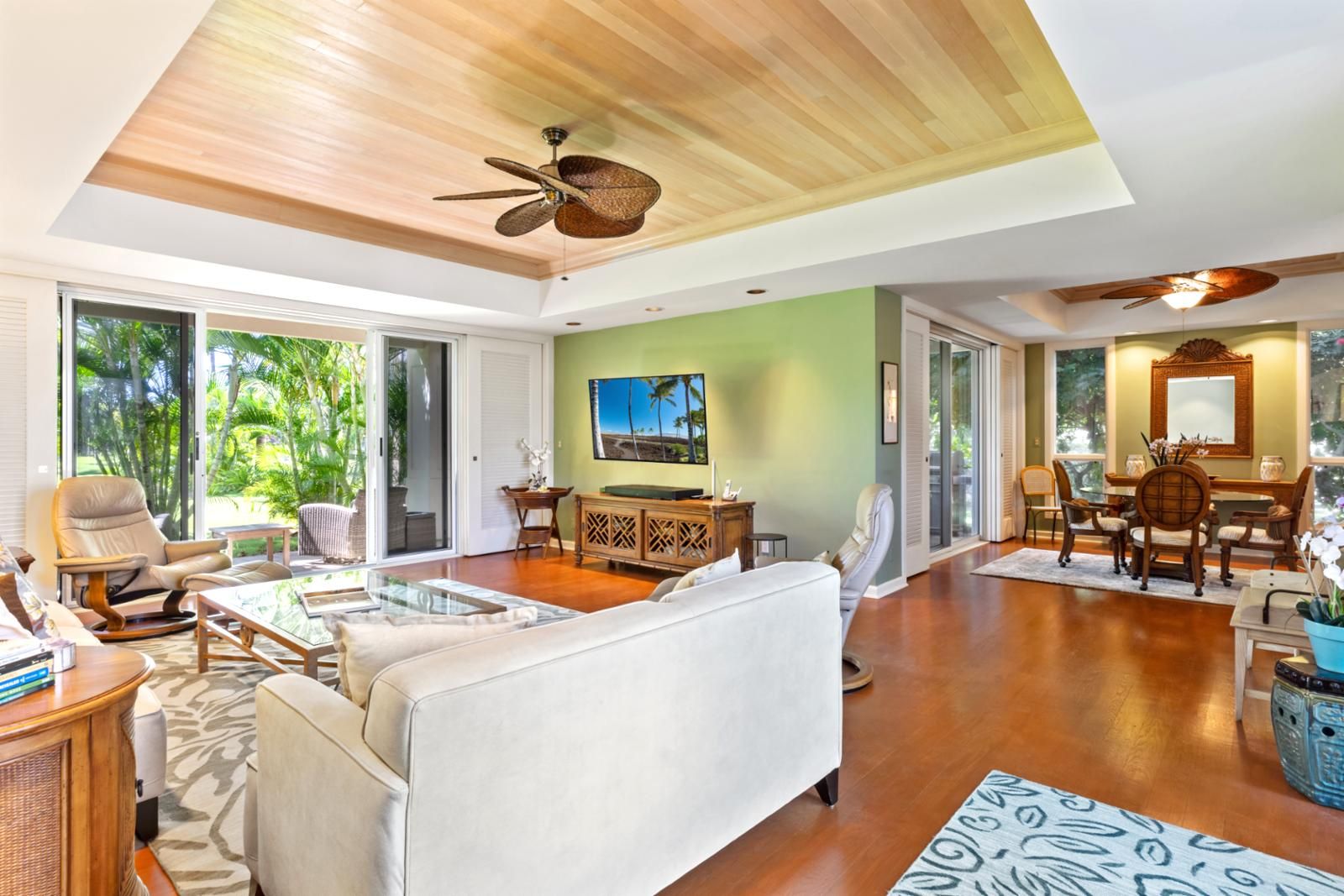 A spacious living room with a large sofa, a coffee table, and a TV. There is a dining area with a table and chairs. The room has wooden flooring and a ceiling fan.