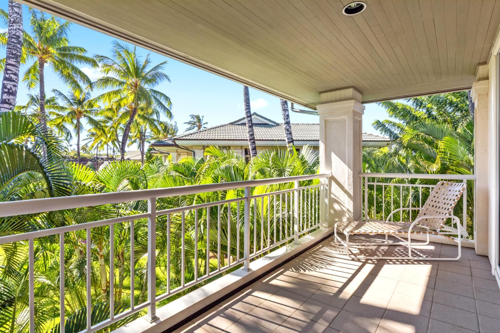 A balcony with a white railing, two wicker lounge chairs, and a view of palm trees and a house in the background.