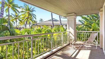 A balcony with a white railing, two wicker lounge chairs, and a view of palm trees and a house in the background.