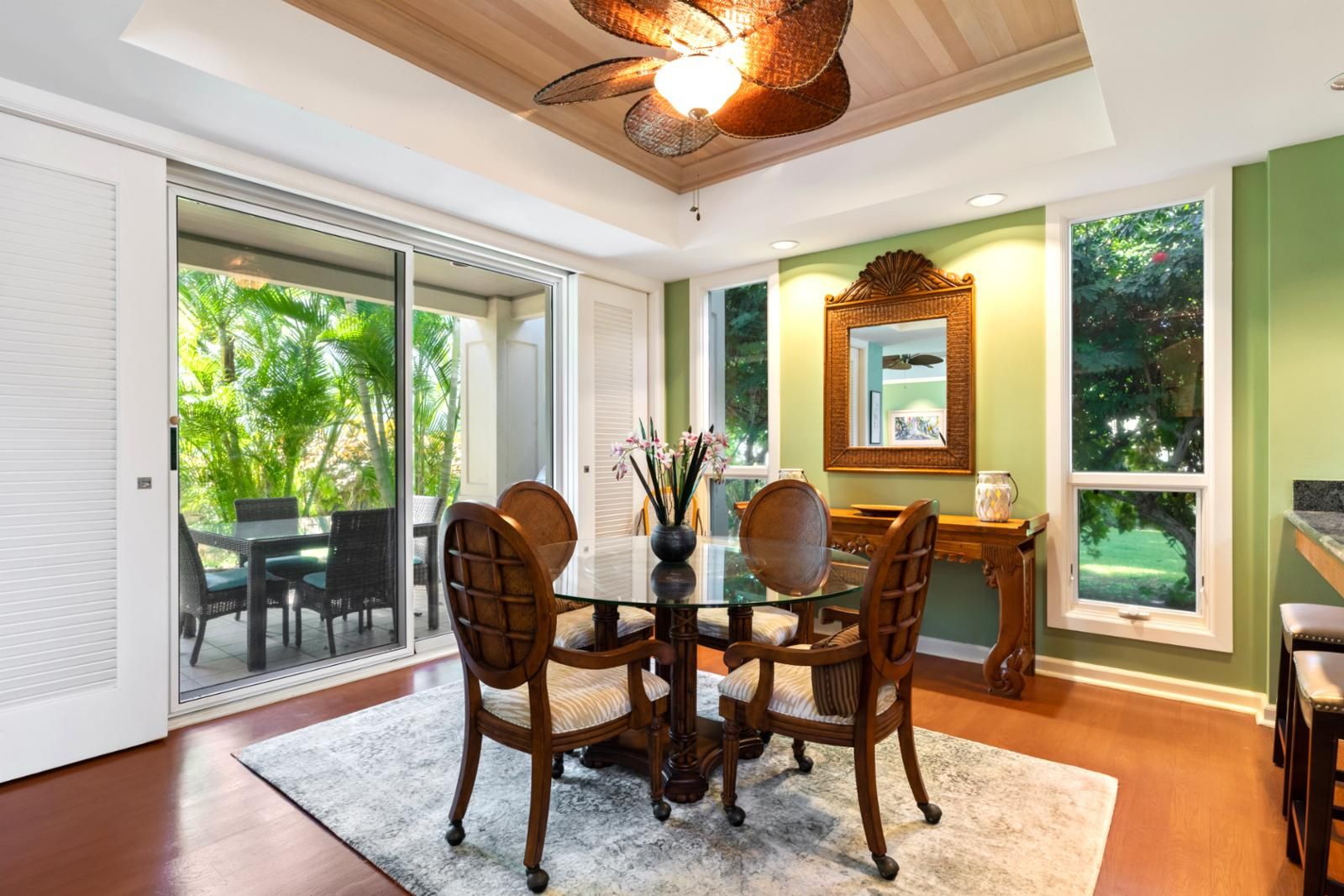 A dining area with a glass table, four chairs, a vase with flowers, and a wooden ceiling fan.