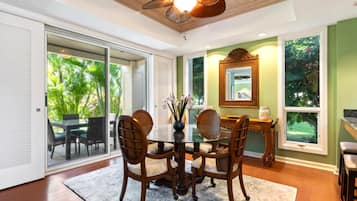 A dining area with a glass table, four chairs, a vase with flowers, and a wooden ceiling fan.