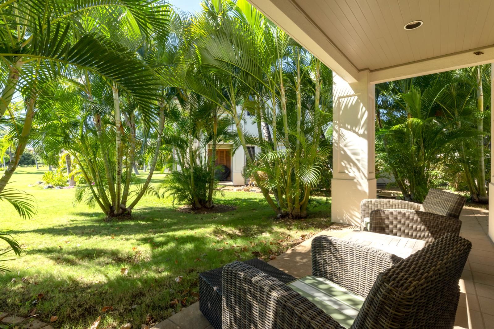 A covered outdoor seating area with wicker furniture, surrounded by lush greenery and palm trees.