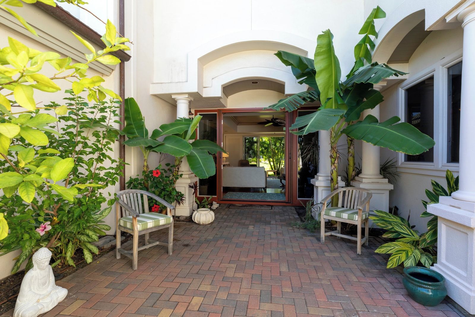 Un patio con piso de ladrillo, dos sillas de mimbre, una estatua y diversas plantas.