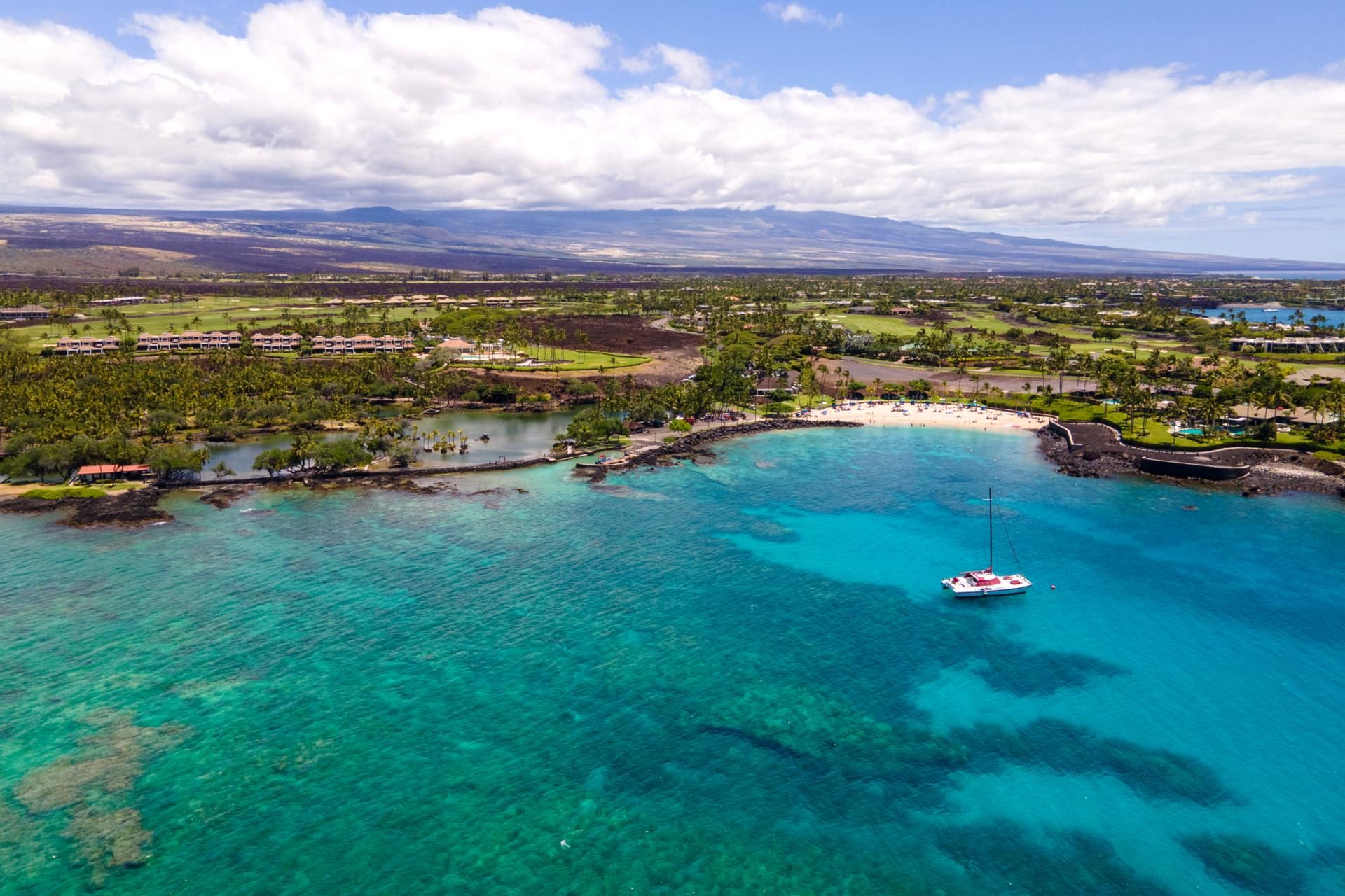 An aerial view of a coastal resort with clear turquoise waters, a sandy beach, and a golf course in the background.