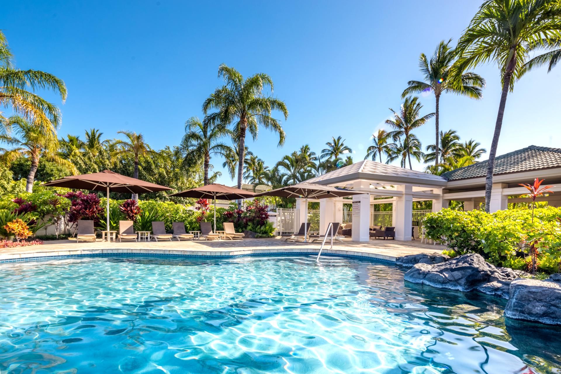 A pool area with lounge chairs and umbrellas, surrounded by palm trees and a building with a tiled roof.