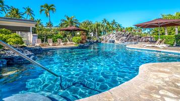 A clear blue pool with lounge chairs and umbrellas, surrounded by palm trees and a waterfall feature.