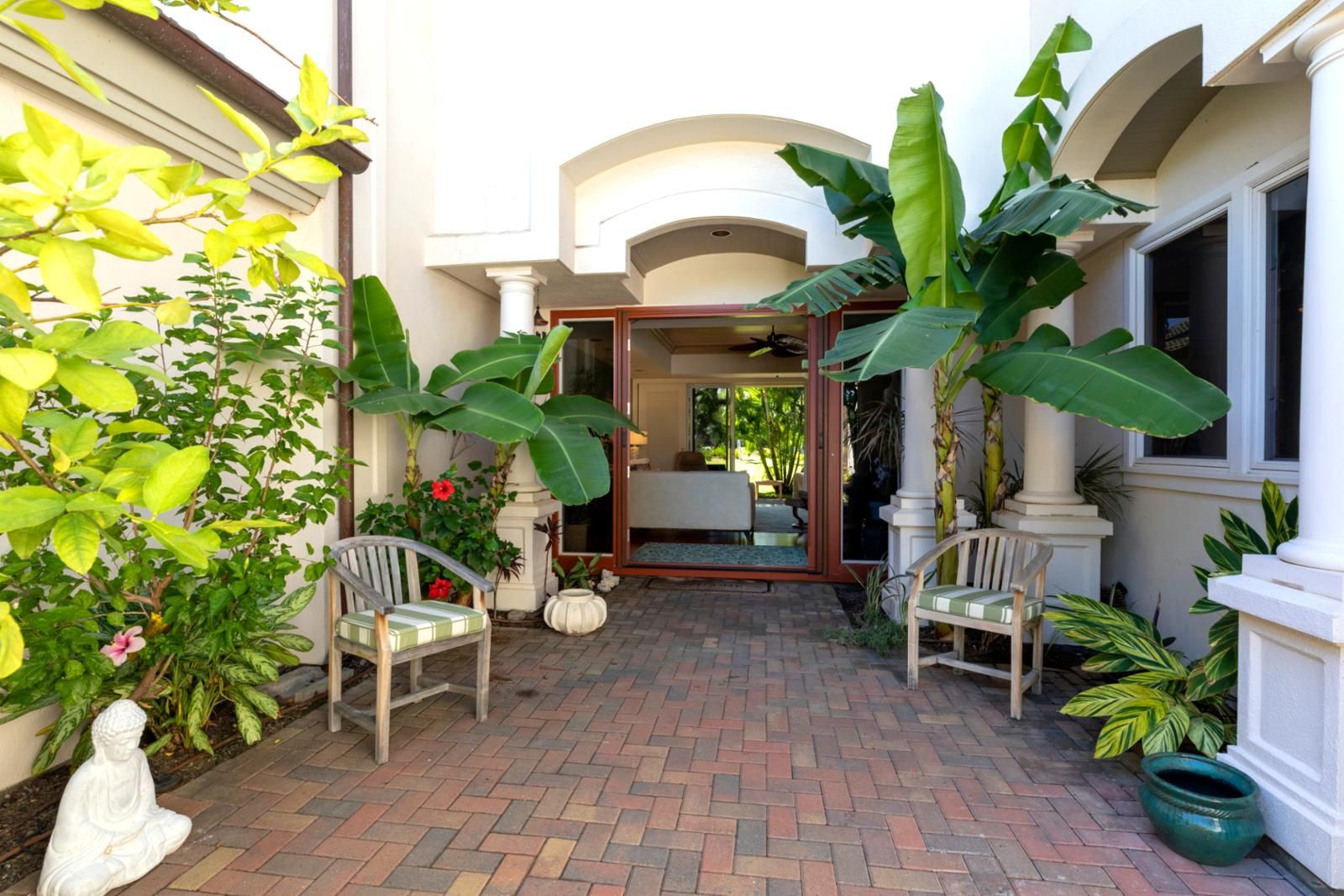A courtyard with a brick floor, two wicker chairs, and large green plants.