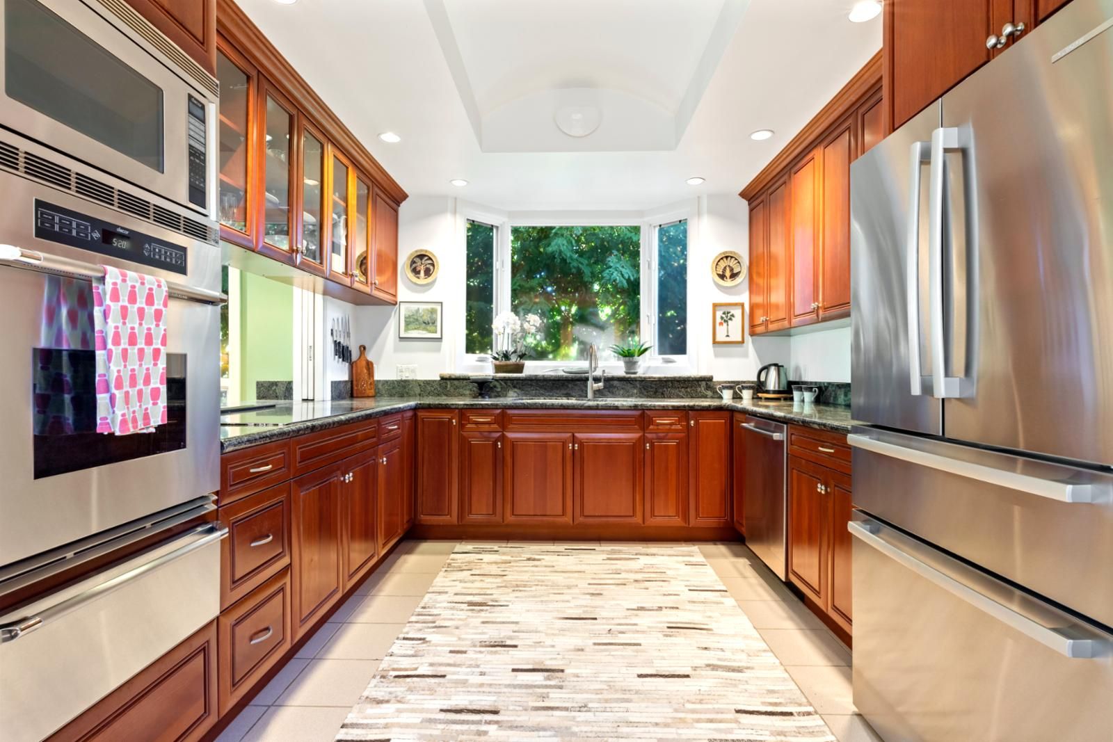 A modern kitchen with wooden cabinets, stainless steel appliances, and a large window offering a view of greenery.
