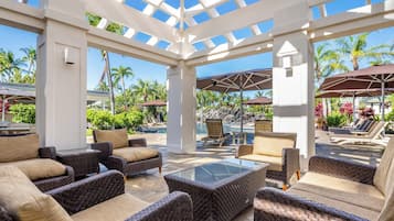 A covered outdoor seating area with wicker furniture and a glass table, overlooking a pool and tropical landscaping.