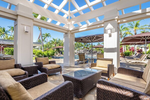 Un espace extérieur aménagé, abrité et doté de mobilier en osier et d’une table en verre, offrant une vue sur une piscine et un jardin tropical.