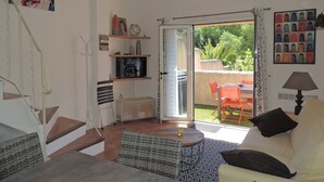 Una sala de estar con un sofá, un televisor, una escalera y un ventanal corredero que da a un área exterior con una mesa y sillas.
