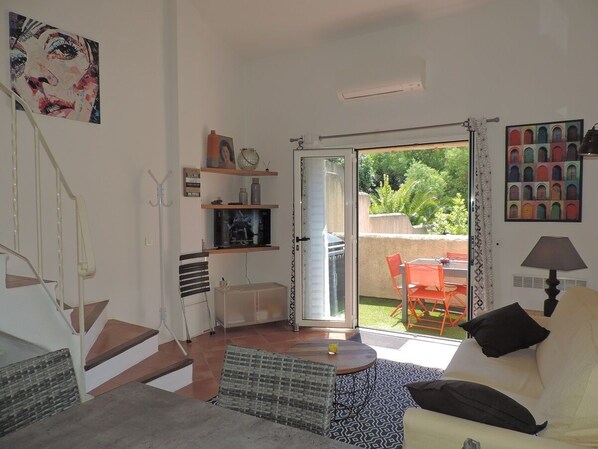 Un salon comprenant un canapé, un téléviseur, un escalier et une porte-fenêtre coulissante donnant sur un espace extérieur avec une table et des chaises.