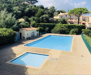 A rectangular swimming pool with a tiled deck, surrounded by greenery and a building in the background.