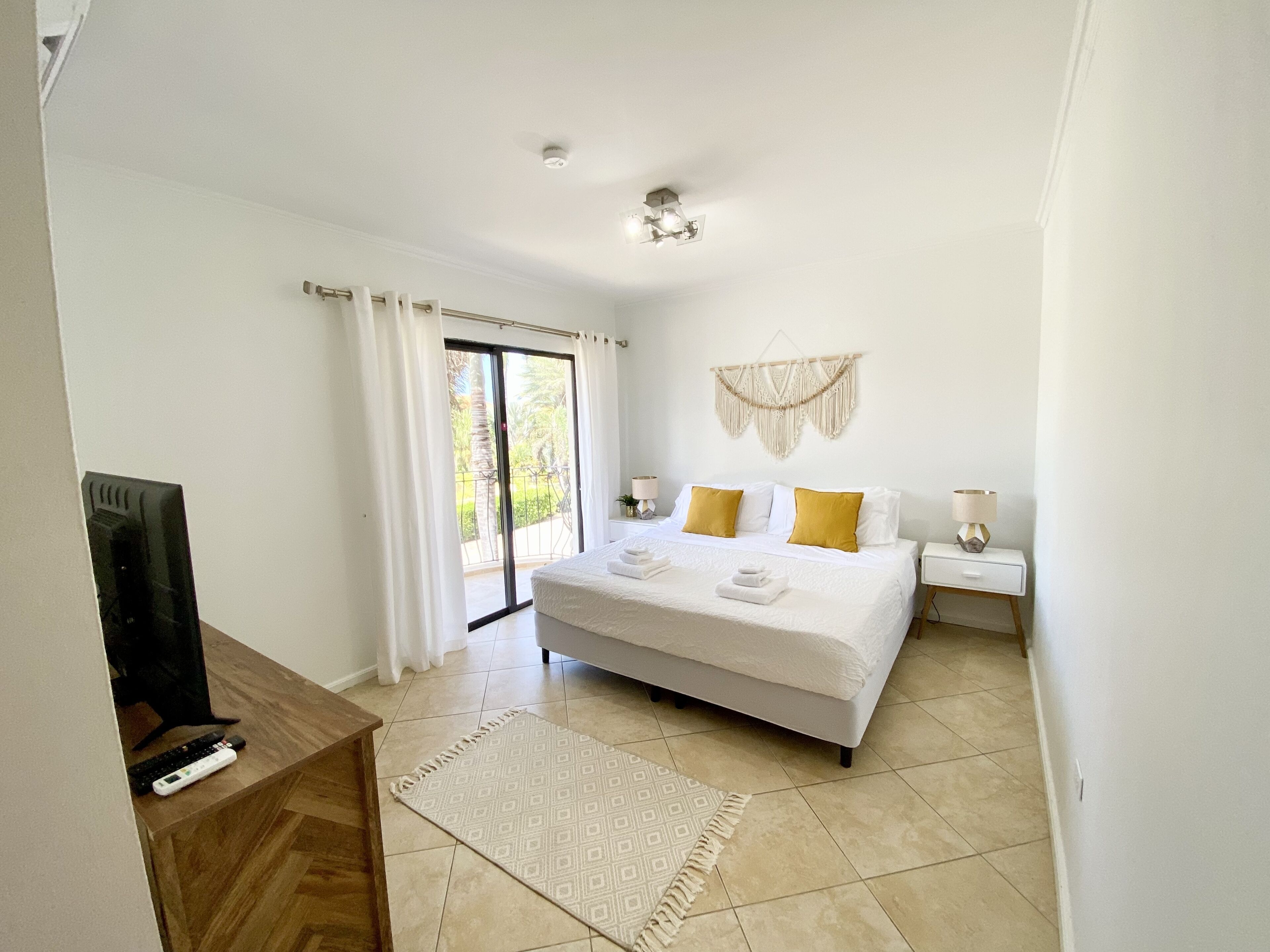 A bedroom with a large bed, a desk with a TV, a sliding glass door to the outside, and a macrame wall hanging.