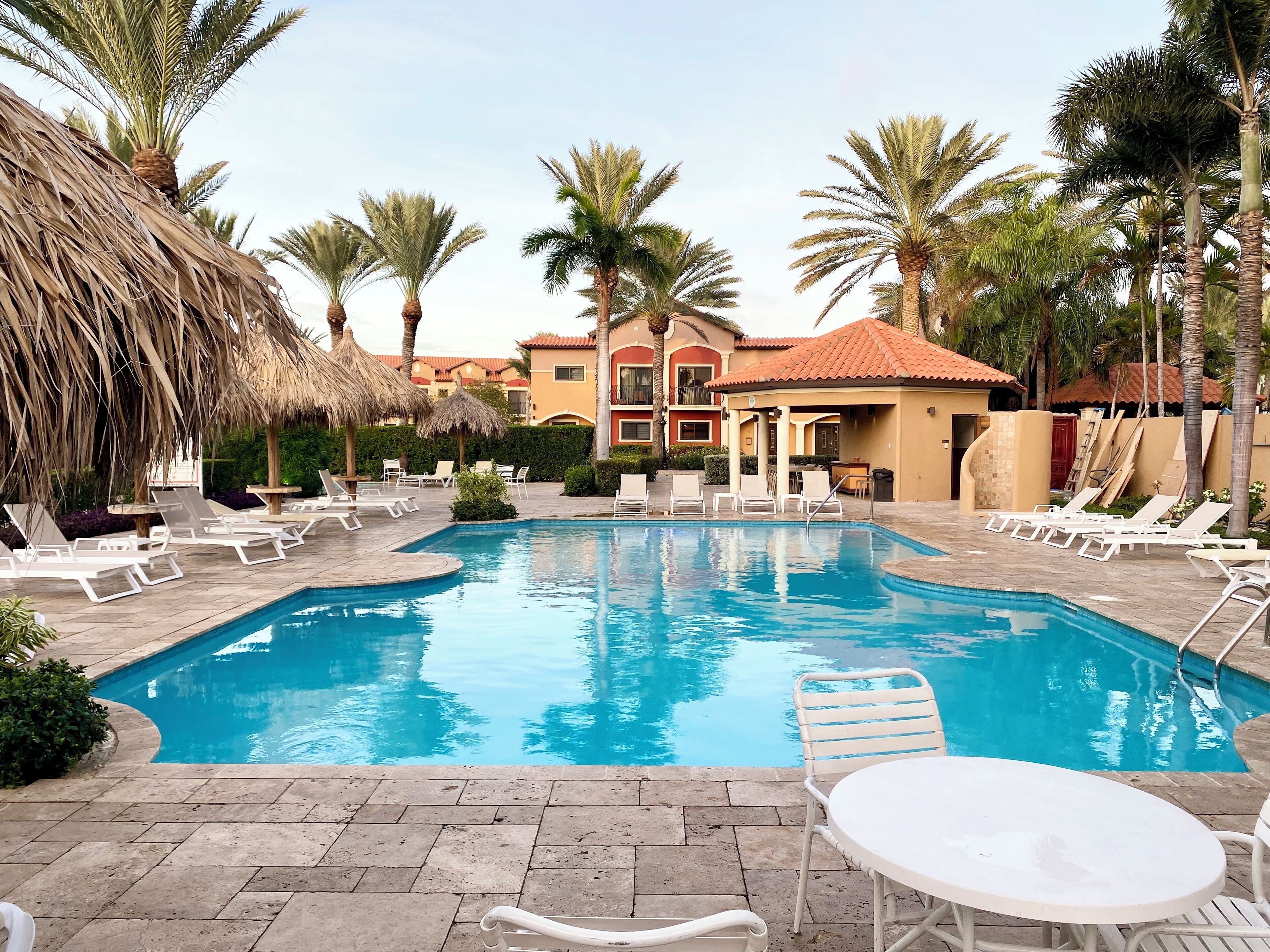 A swimming pool with lounge chairs and a table, surrounded by palm trees and buildings.