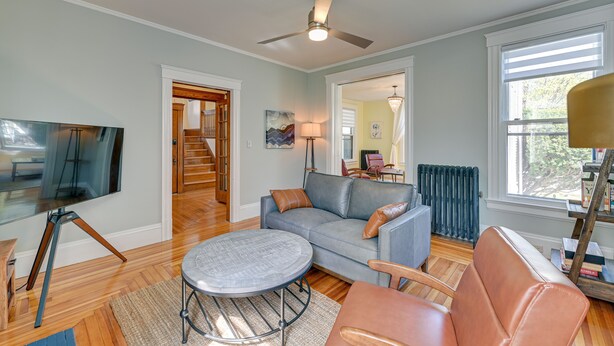 A living room with a grey sofa, a round coffee table, a TV on a tripod stand, and a ceiling fan. - Spacious Historic Home in the Berkshires! (Pittsfield)