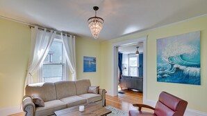 A living room with a beige sofa, a wooden coffee table, a red leather chair, a chandelier, and a large wave painting on the wall. - Spacious Historic Home in the Berkshires! (Pittsfield)
