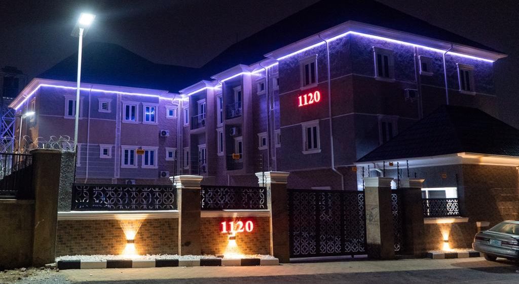 Un immeuble à plusieurs étages, dont l’entrée est marquée par une enseigne bien visible portant le numéro « 1120 », et qui est éclairée par des projecteurs au sol.