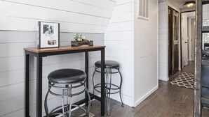 A kitchen with white wooden walls, a wooden table with two black stools, and a framed photo on the table. - Tiny AFrame/Fire Pit/Pets/Lake Access/Dogwood Canyon (Lampe)
