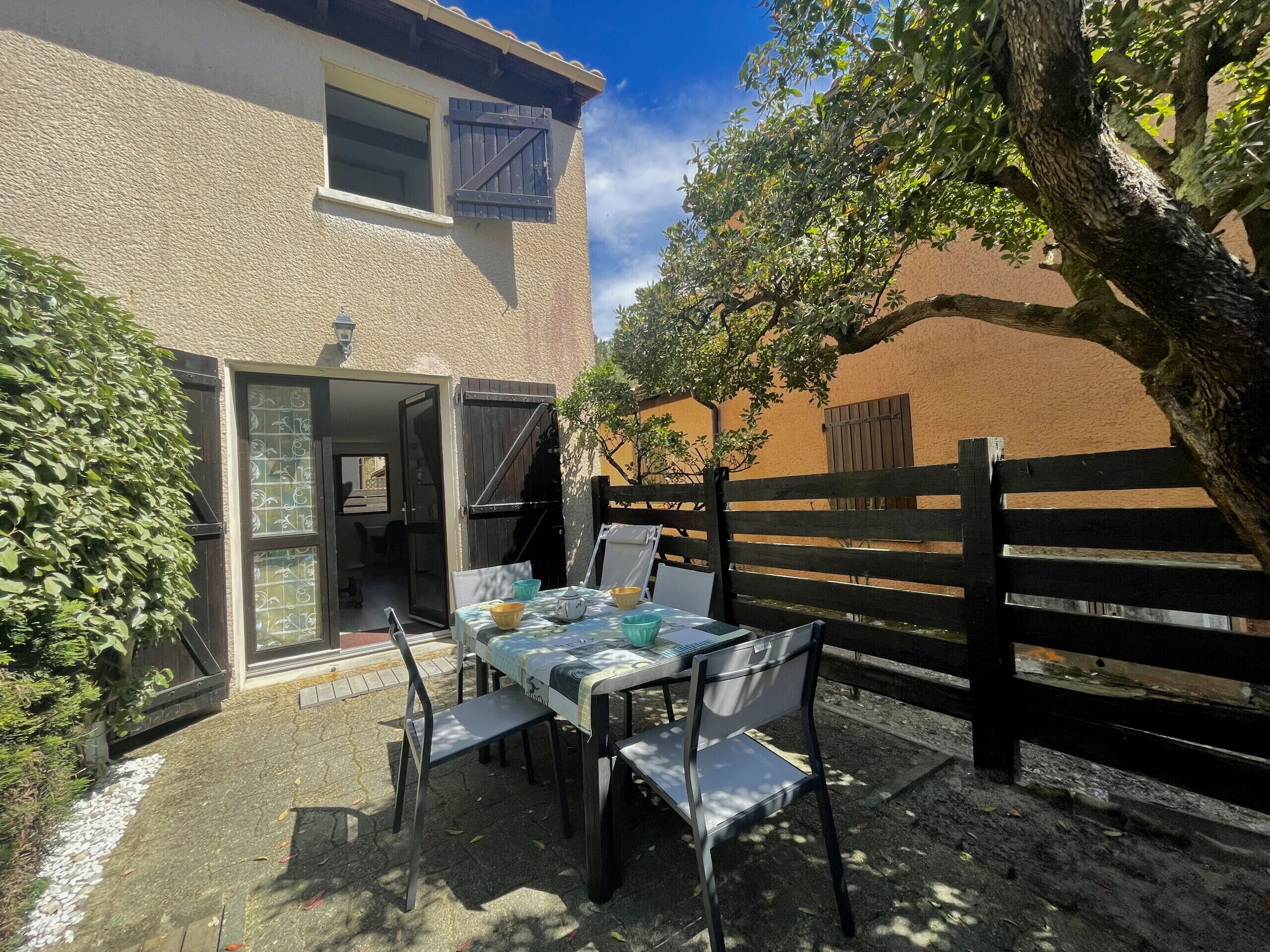 Een terras buiten met een tafel en stoelen, een houten omheining en een gebouw met een raam en een deur.