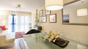 A modern hotel room with a glass dining table, a sofa, and a large window with curtains.