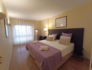 Habitación de hotel con una cama grande, un escritorio, una silla, una lámpara y un cuadro en la pared.