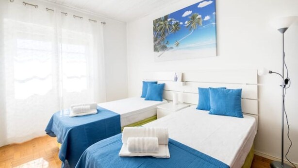 Habitación de hotel con dos camas, un cuadro de palmeras, una lámpara de pie y una ventana con cortinas translúcidas.