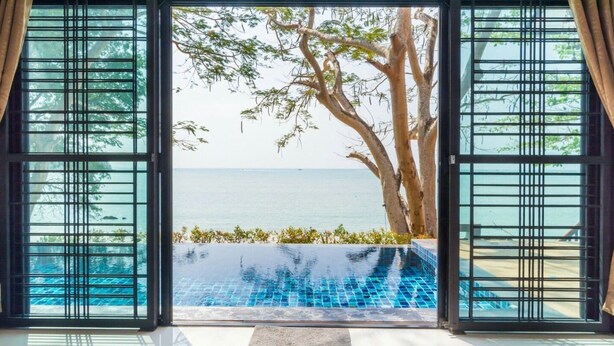 Vista a través de un vidrio corredero a una piscina con una vista despejada del mar y un árbol.