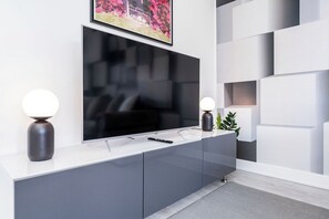 Uma sala de estar moderna com uma grande televisão de tela plana presa a uma parede branca, uma mesa console cinza elegante com duas luminárias esféricas e um quadro emoldurado acima da TV.