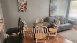 A living room with a grey sofa, a glass coffee table, and white wooden chairs. - Derby rental furnished 2BR, 1 full bath(shower/tub) house (Louisville)