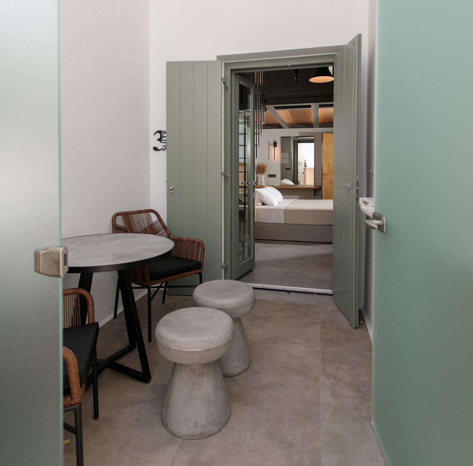 A compact room with a round table, two chairs, and two stools. A door leads to a bedroom with a bed and a mirror.