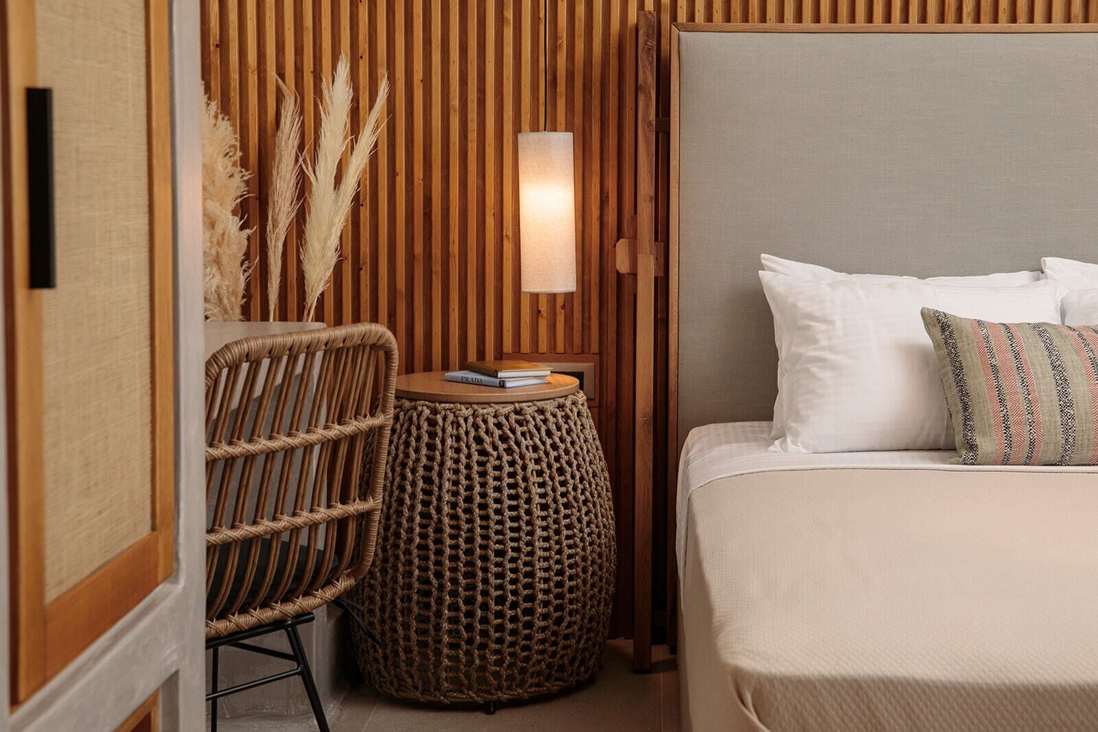 A bedroom with a bed, a wicker chair, a round woven side table, and a wall-mounted lamp.