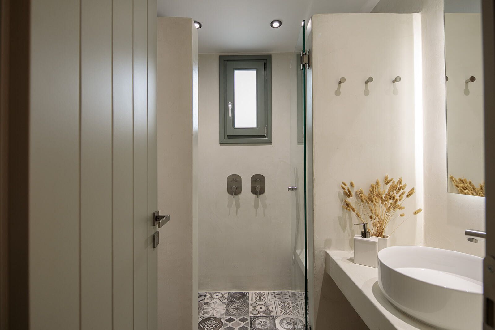 A modern bathroom with a glass shower enclosure, a small window, and a white sink with a vase of dried flowers.