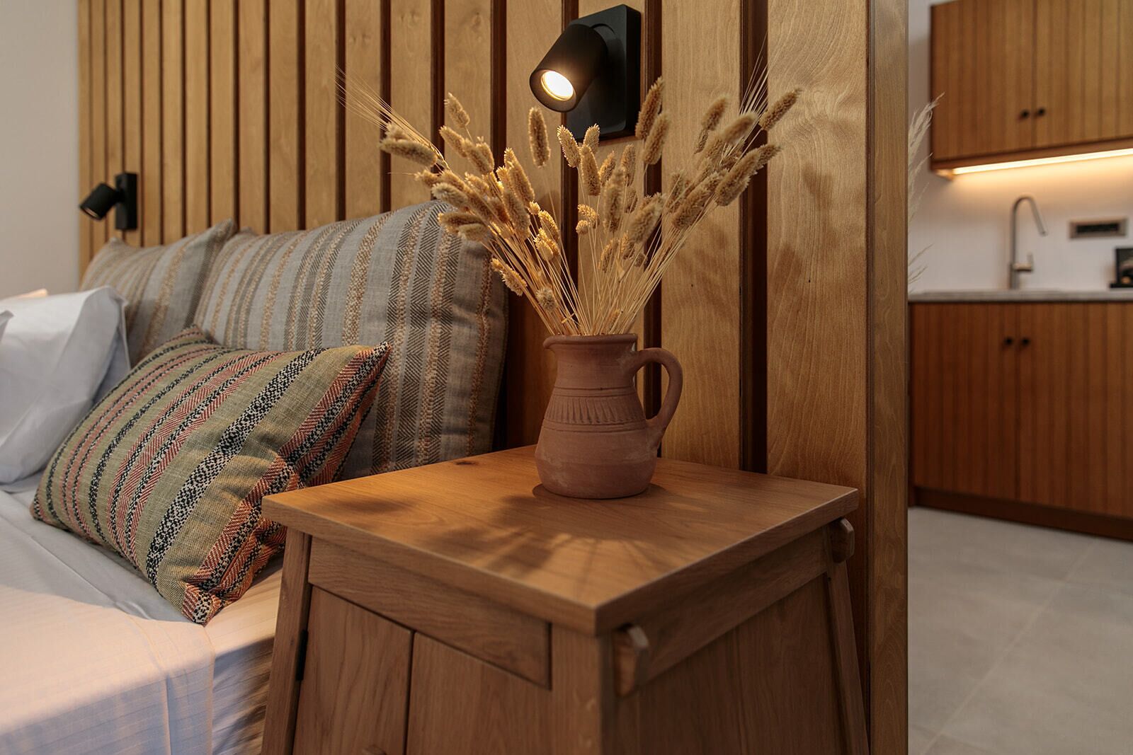 A wooden bedside table with a vase of dried wheat stalks, a patterned pillow, and a wall-mounted light.