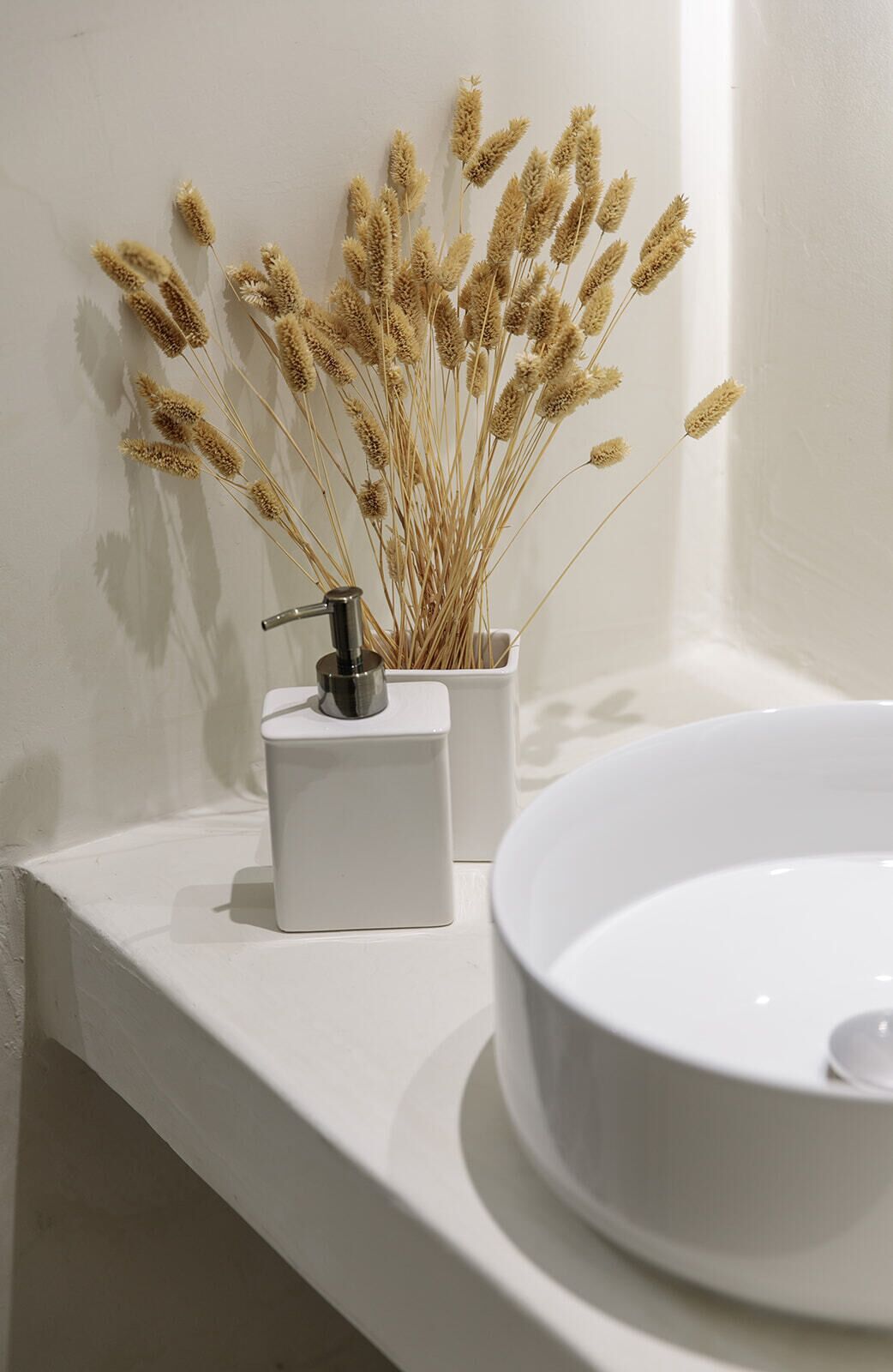 Un baño con un lavamanos blanco y una pequeña maceta blanca con flores secas.