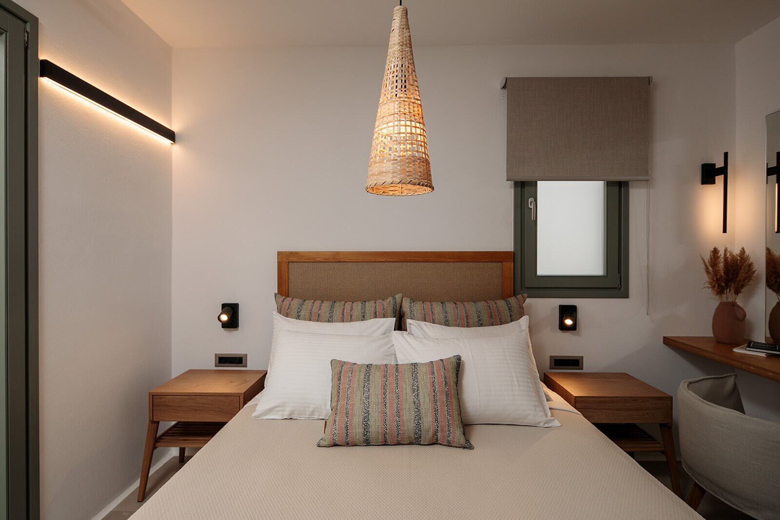 A modern bedroom with a large bed, two bedside tables, a hanging light fixture, a window with a blind, and a mirror on the wall.