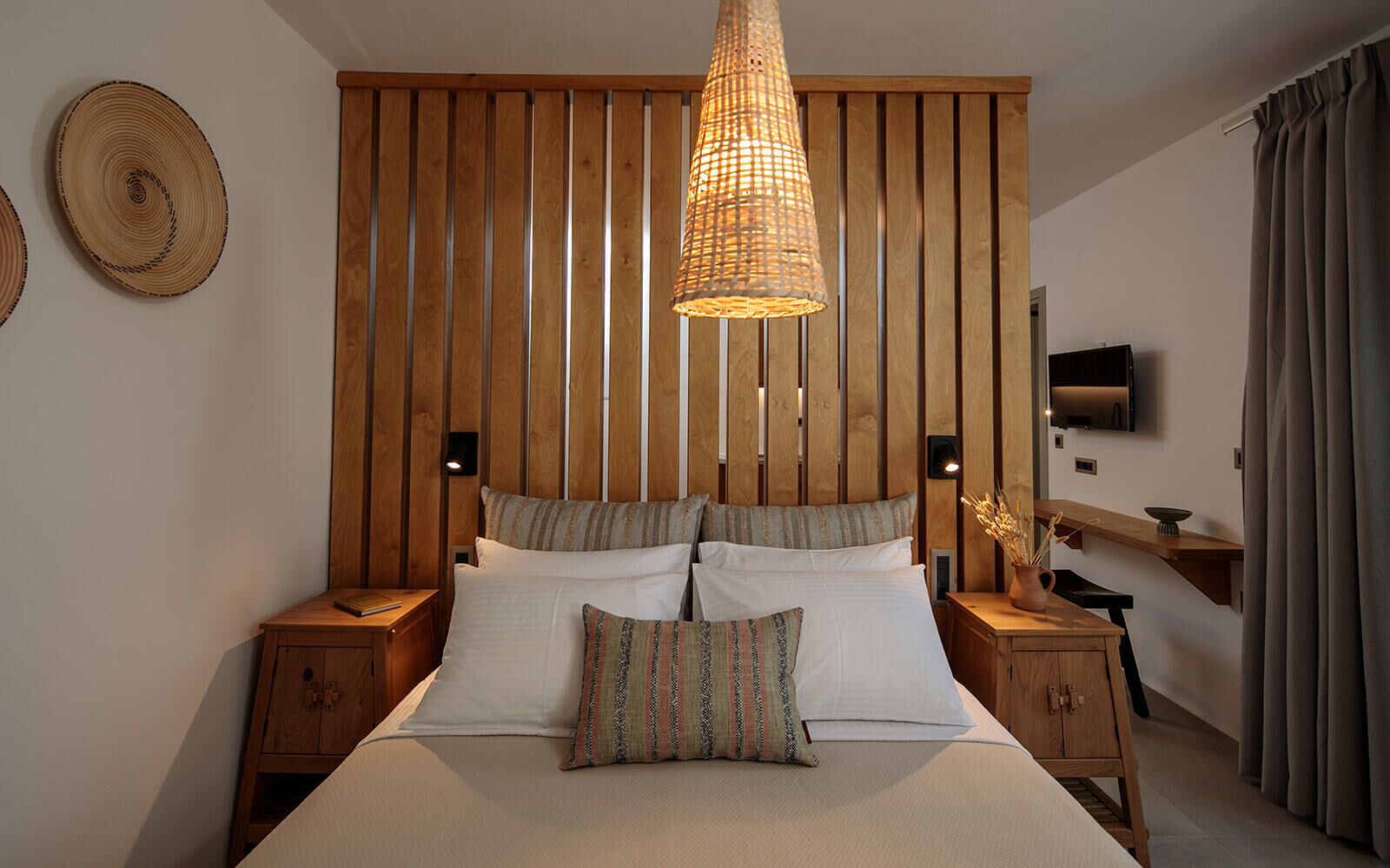 Un dormitorio con cabecera de listones de madera, una cama grande con muchos cojines, mesitas de noche, un televisor y un pequeño armario de madera.
