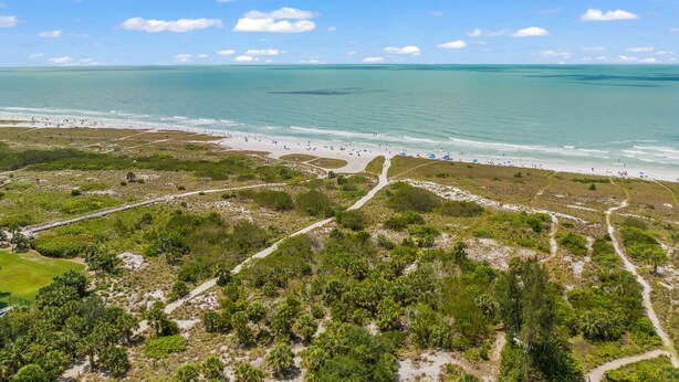 An aerial view of a beach with clear skies, a sandy shore, and a mix of greenery and paths.