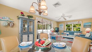 A dining area with a glass table, chairs, and a buffet bowl. There is a sofa and a ceiling fan in the background.