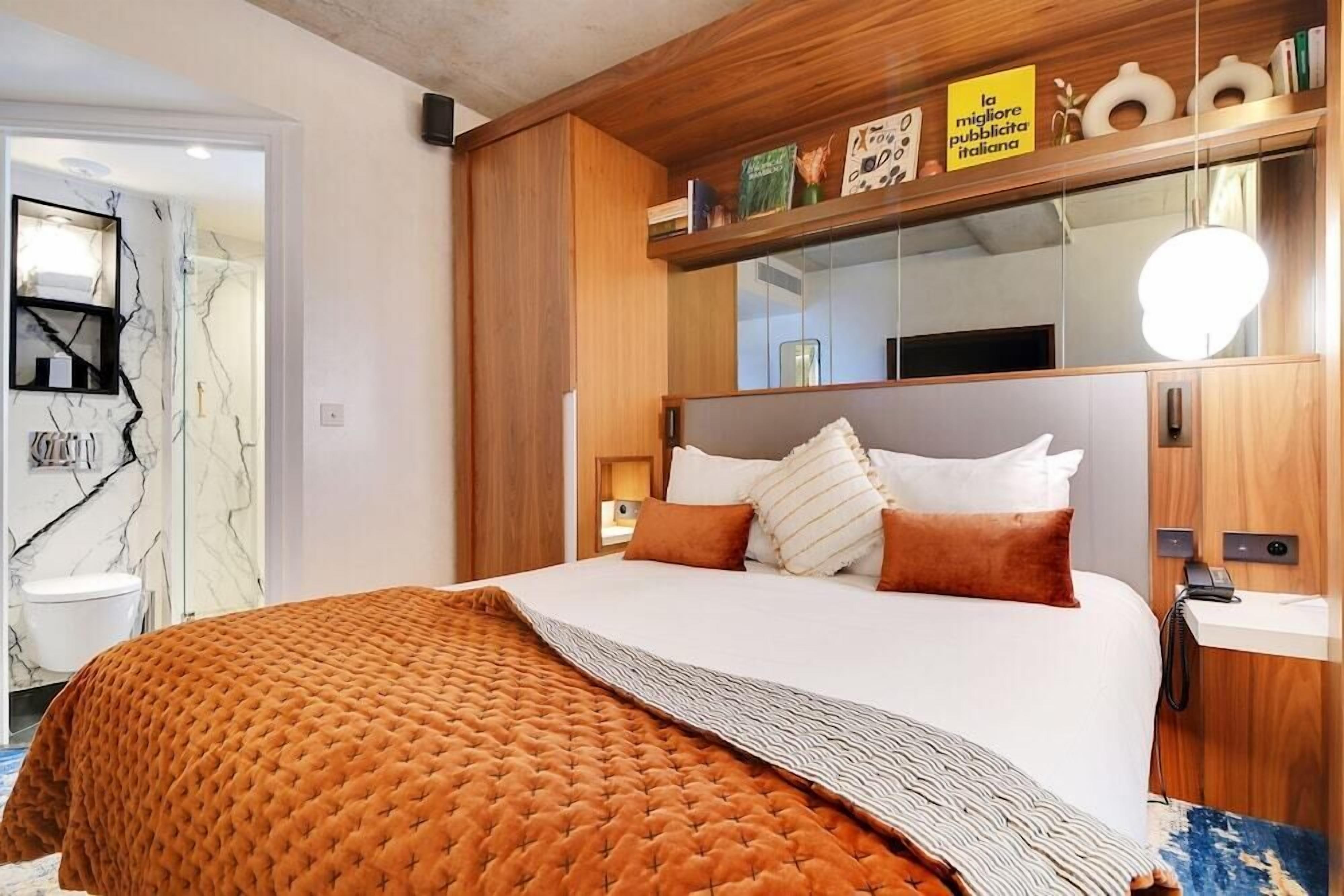 A hotel room with a large bed, a wooden headboard, a bedside table, a mirror, and a shelf with books and decorative items.