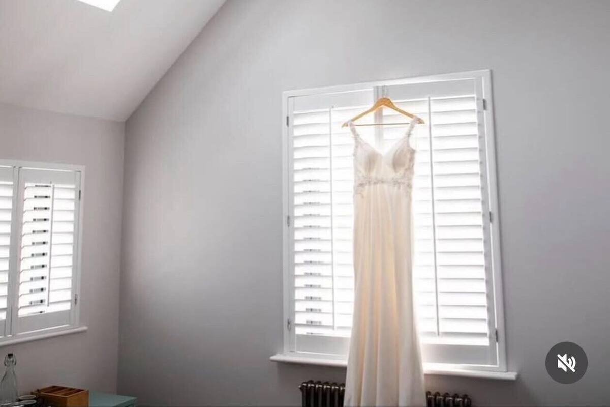 A room with a sloped ceiling, a hanging light fixture, a window with blinds, and a dress on a hanger.
