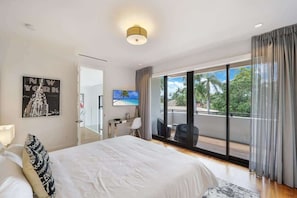 Ein modernes Hotelzimmer mit einem großen Bett, einem Schreibtisch, einem Fernseher und einem Balkon mit Blick auf Palmen.