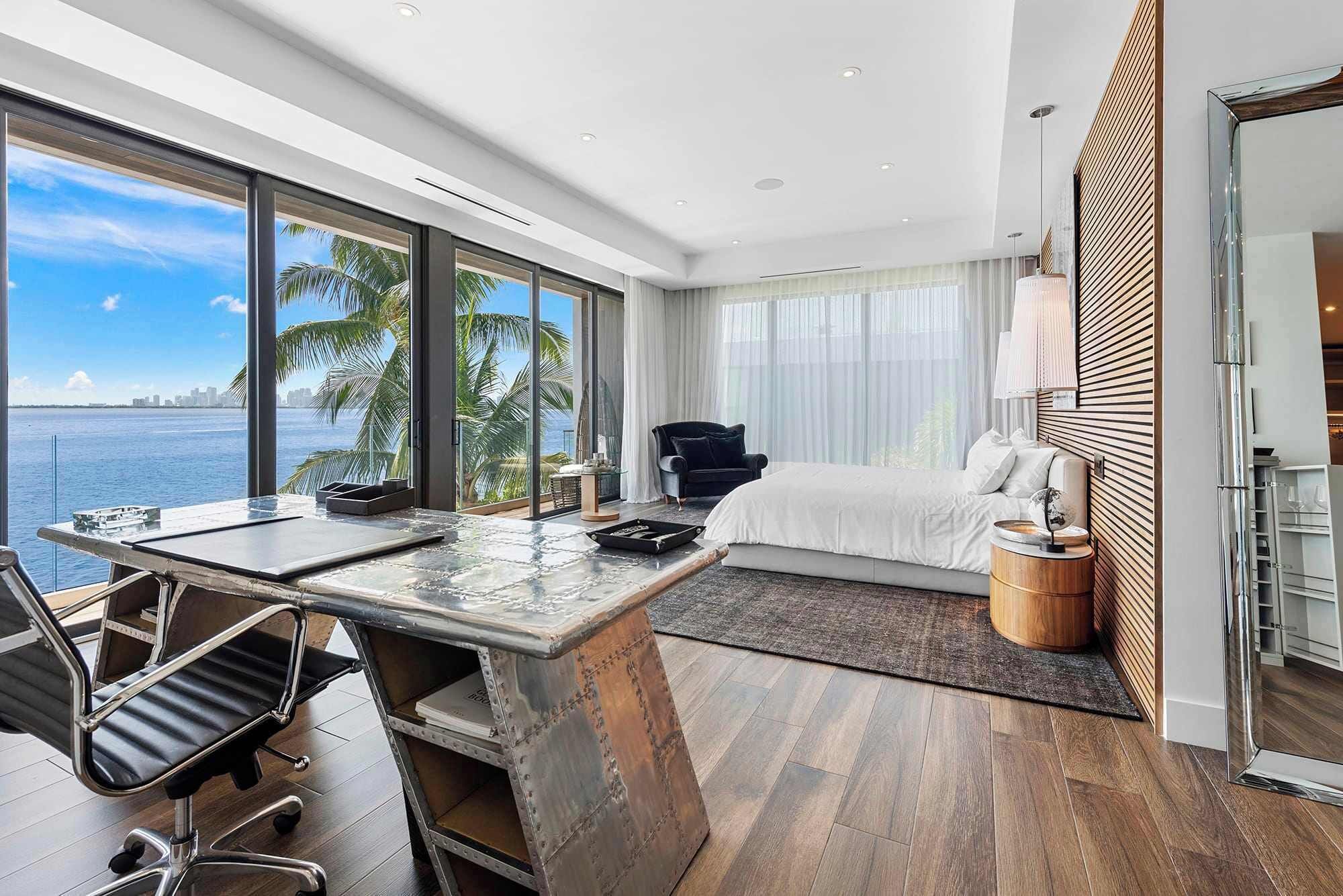 A modern hotel room with a large window overlooking the sea, a bed, a desk with a chair, and a small side table.