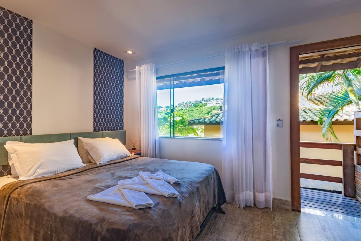 Une chambre d’hôtel avec un lit, des oreillers blancs, une fenêtre avec des rideaux et une vue sur la verdure à l’extérieur.