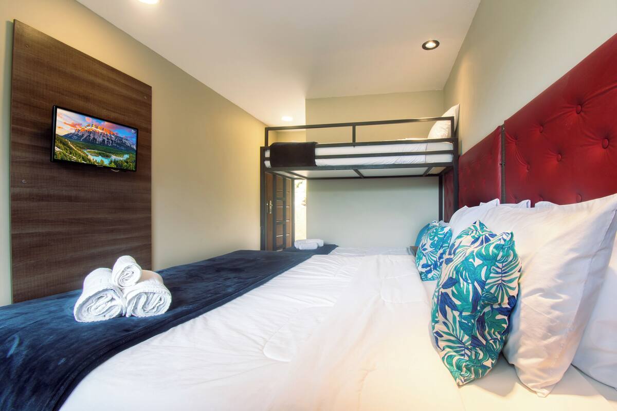Une chambre d’hôtel avec un lit superposé, un lit simple recouvert d’une couverture bleue et agrémenté d’oreillers blancs, un téléviseur fixé au mur et un tableau représentant des montagnes.