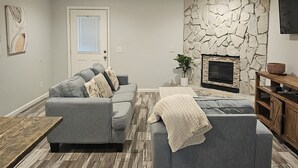 A living room with a grey sofa, a wooden coffee table, a stone wall, a fireplace, and a television mounted on the wall.
