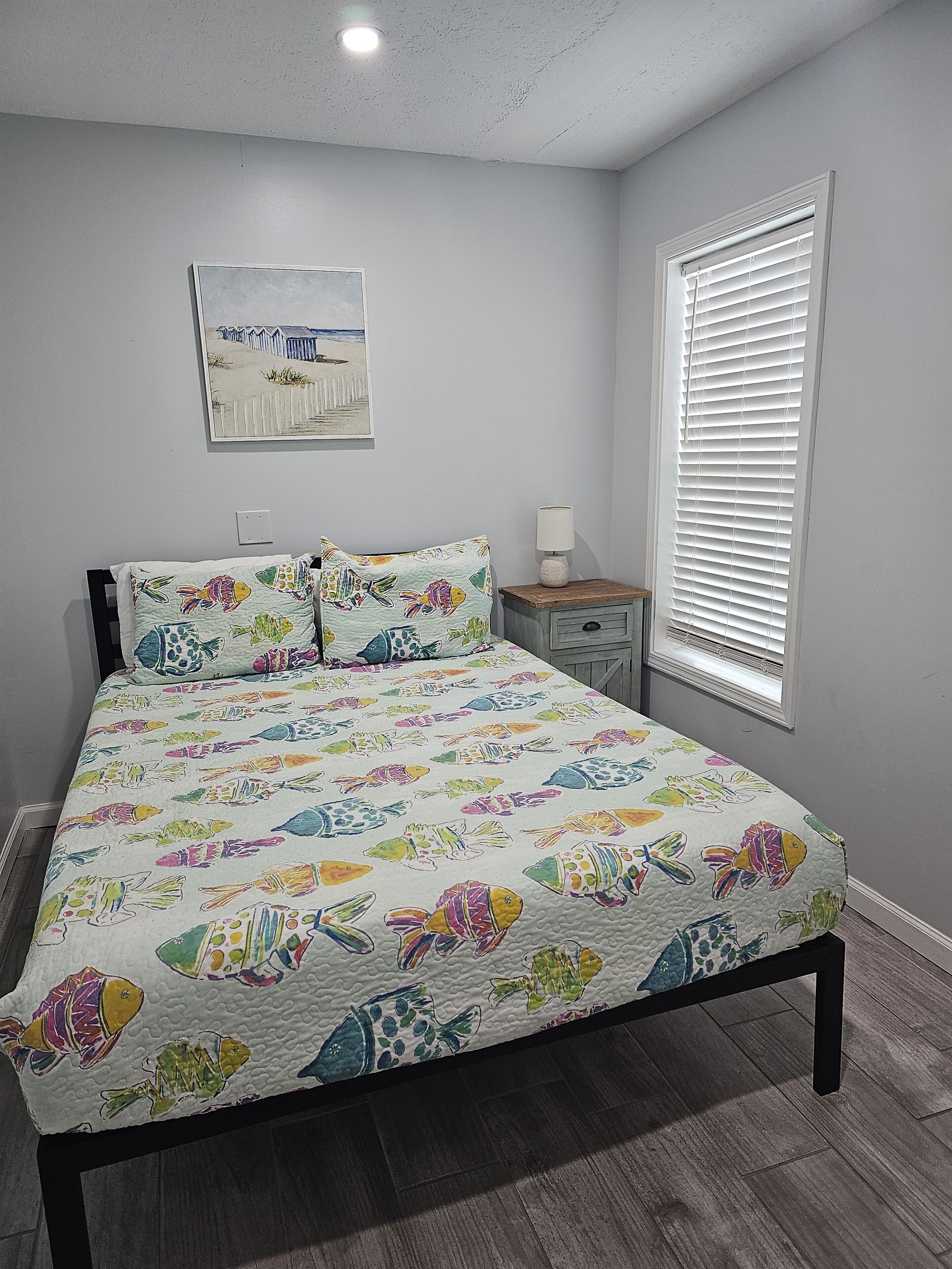 A bedroom with a bed, a nightstand, a lamp, a window with blinds, and a wall-mounted picture.